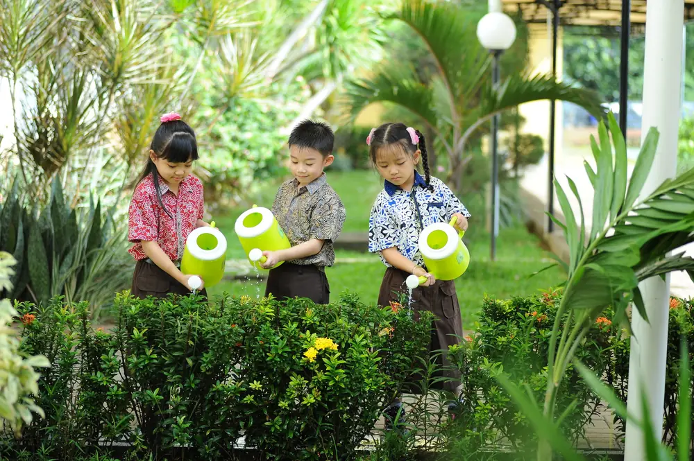 Three young children water plants in a garden with bright green watering cans
