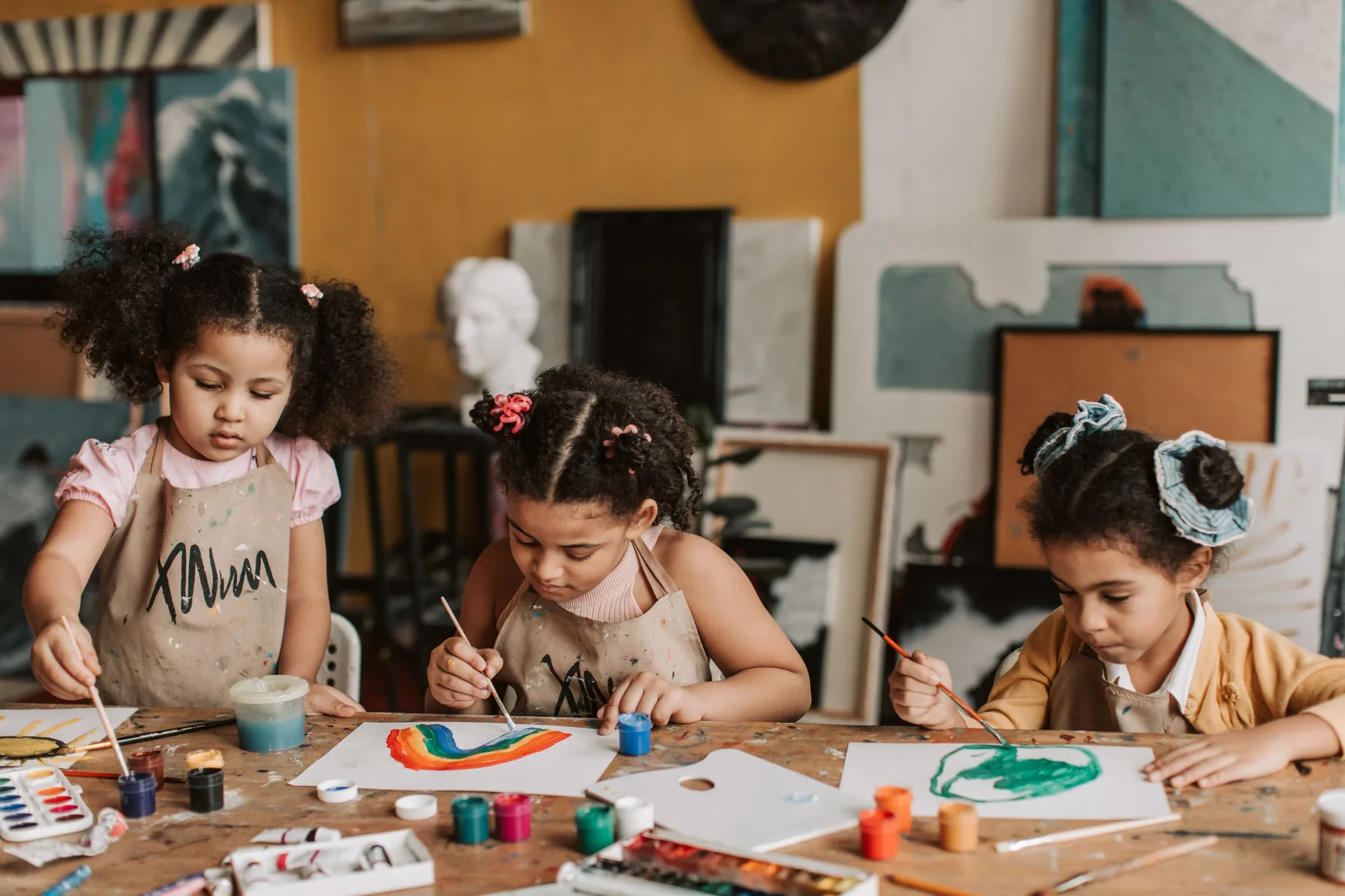 Three young students in paint-spattered aprons painting at a wooden table in the art studio