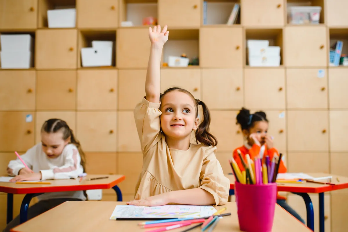 A smiling girl raises her hand at her desk while classmates draw behind her