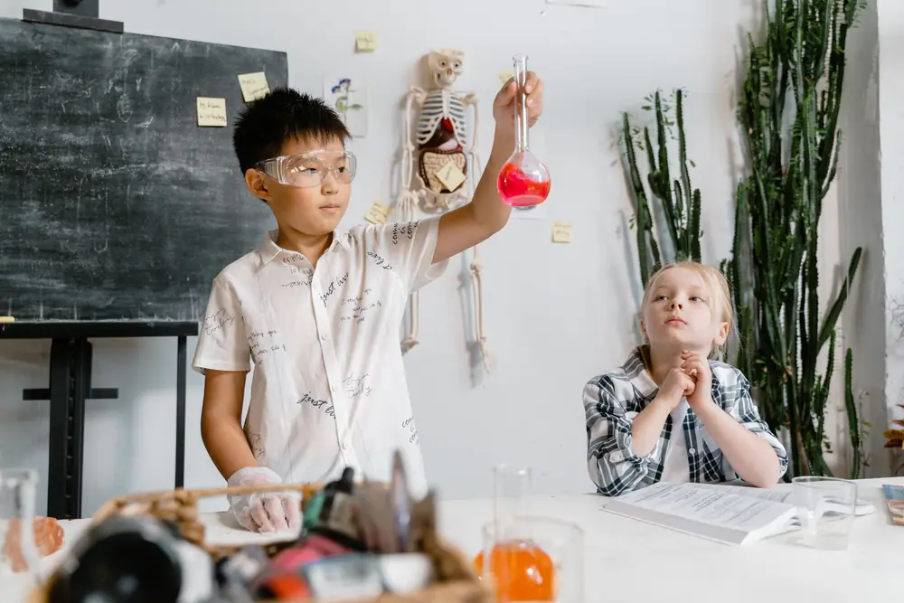 A boy in safety glasses holds up a flask during a science lab while a classmate watches