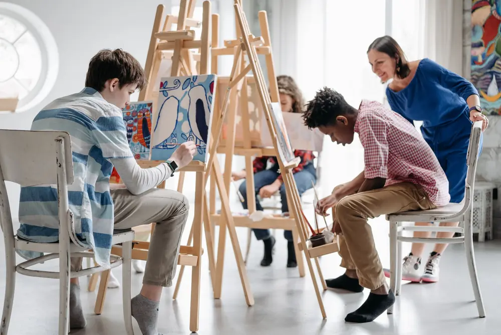 Students painting at wooden easels in a bright art studio while their teacher looks on